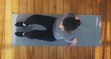 Young plump woman in sportswear and sneakers lies down on mat to do sports exercises on wooden floor upper view slow motion video