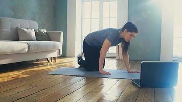 woman with ponytail in sportswear does low plank on mat by laptop put on wooden floor at online training slow motion