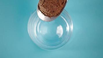 Verter cacao en polvo de una cuchara en un vaso vacío y limpio sobre una mesa azul en el estudio con vista cercana desde arriba a cámara lenta