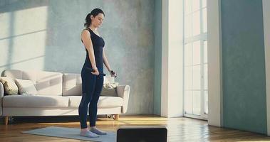 Tranquil young woman in stylish sportswear does exercise for arms with dumbbell bars near window in sunny living room slow motion