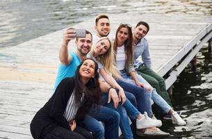 A group of young people sitting on the edge of the pier, and make selfie. Friends enjoying a game on the lake photo