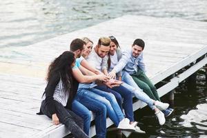 Portrait of joyful group of young people sitting on the edge of the pier, outdoors in nature. Friends enjoying a day on the lake. photo
