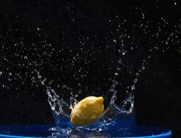 limón amarillo cayendo en el agua azul sobre un fondo negro foto