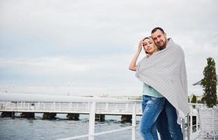 Happy young couple in a blanket, standing on the pier in the water. photo