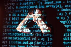 Double exposure of a caucasian man and Virtual reality headset is presumably a gamer or a hacker cracking the code into a secure network or server, with lines of code photo