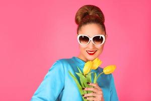 retrato de una joven hermosa mujer vestida de seda azul y gafas de sol blancas con tulipanes amarillos en el fondo rosa foto