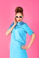 retrato de una joven hermosa mujer con vestido de seda azul y gafas de sol blancas de fondo rosa foto