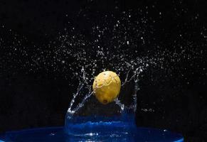 limón amarillo cayendo en el agua azul sobre un fondo negro foto