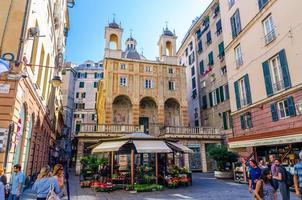 Genoa, Italy, September 11, 2018 people are walking down square with small church Chiesa di San Pietro in Banchi piazza and flower stall kiosk among buildings in historical city Genova centre