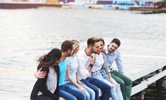 Portrait of joyful group of young people sitting on the edge of the pier, outdoors in nature. Friends enjoying a day on the lake. photo