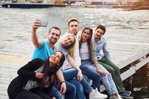 A group of young people sitting on the edge of the pier, and make selfie. Friends enjoying a game on the lake
