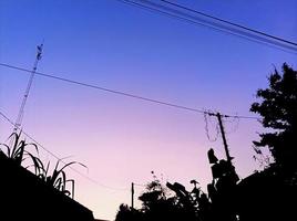 foto de silueta de casas, postes de electricidad y plantas al amanecer en el cielo azul, violeta y rosa del pueblo.