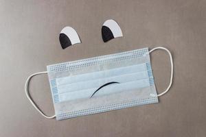 Blue disposable medical mask with sad smiley face with drawn eyes and mouth on a gray background. With copy space photo