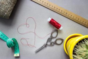Sewing accessories scissors, measuring tape, ruler, fabric. In center of composition - red thread, laid out in the shape of a heart, with a needle at the end. With cactus for home furnishings