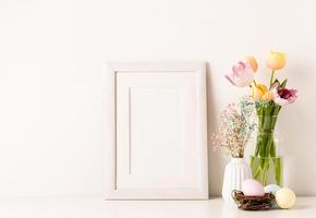 Mockup with a white frame spring tulips in vase, gypsophila and pastel colored eggs on light background photo