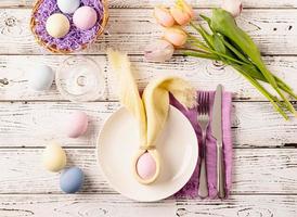 Table setting for Easter feast. Easter eggs, bunny, tulips and cutlery top view flat lay on white rustic background photo