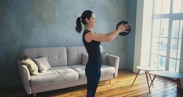 Brunette young woman in black tracksuit does sports exercises with weight plate near sofa in living room at home slow motion video