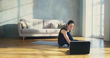 attraktiv kvinna i sportkläder trycker på tangenten på den bärbara datorn och lägger sig på mattan vid onlineträning i soligt vardagsrum i slow motion video