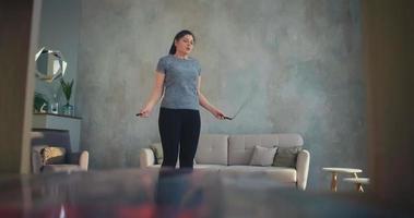 dama elegante en camiseta salta entrenamiento de cuerda contra fondos de pantalla diseñados y sofá en la vista de la sala de estar desde la mesa a cámara lenta video