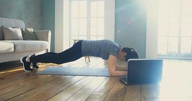 sterke jonge vrouw in trainingspak staat in plank pose kijken naar video op laptop in woonkamer bij quarantaine isolatie slow motion