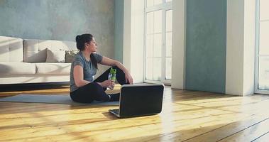 Pretty tired young woman drinks water resting after intensive sports training near modern laptop in living room slow motion video