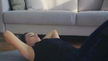 Strong young woman in dark tracksuit does crunches on grey mat against grey sofa in spacious living room closeup slow motion video