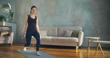 Concentrated young woman in tracksuit does dynamic lunges holding weights training on mat in living room at home slow motion video