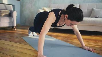 Una deportista fuerte con un chándal cómodo hace entrenamiento de flexiones en una alfombra gris en una espaciosa sala de estar en casa a cámara lenta