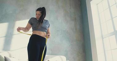 Worried brunette woman in top and leggings measures waist with tape and becomes sad in living room low angle shot slow motion video