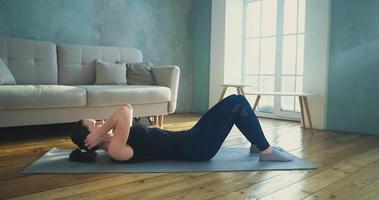 Positive young woman in dark sportswear does crunches lying on grey mat near modern sofa in spacious living room slow motion video