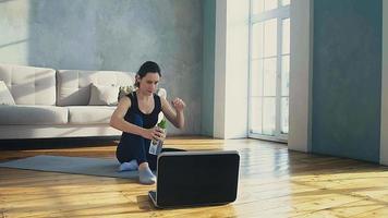 mujer joven con una botella de agua limpia la frente descansando después de entrenar y viendo videos en una laptop en una espaciosa habitación soleada a cámara lenta