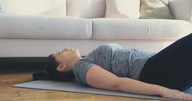 Athletic young woman in tracksuit does crunches and leg lifts lying on grey mat in light living room close view slow motion video