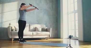 Concentrated young woman in tracksuit does sports exercises lifting dumbbell bars near phone on tripod in living room slow motion video
