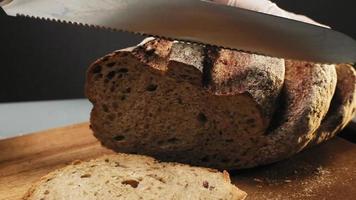 Person in gloves cuts slice of delicious rye bread with sharp knife on wooden board on black background closeup slow motion video