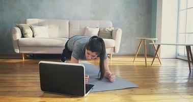 Strong woman in tracksuit stands in plank pose watching video on laptop in living room at quarantine isolation slow motion