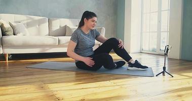 Positive woman in sportswear does crunches and leg lifts near phone on tripod at online training in living room slow motion video