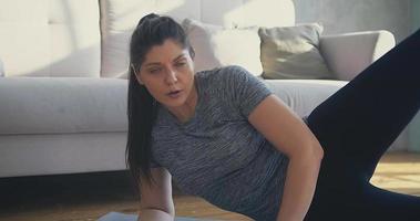 mulher atlética de camiseta cinza e leggings levanta a perna lateral supina no tapete contra o sofá cinza na sala vista de perto em câmera lenta video