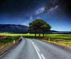 carretera asfaltada y árbol solitario bajo un cielo nocturno estrellado y la vía láctea foto
