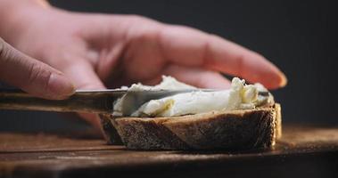 Une femme se répand du fromage frais sur une tranche de pain de seigle avec un couteau brillant sur une planche à découper en bois dans la cuisine vue rapprochée extrême video