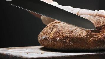 Chef in latex gloves cuts lump of tasty rye bread loaf with knife on wooden board on black background macro slow motion