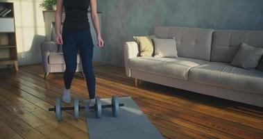 Concentrated sportswoman in black tracksuit takes dumbbells to do exercises near sofa in light living room at home slow motion video