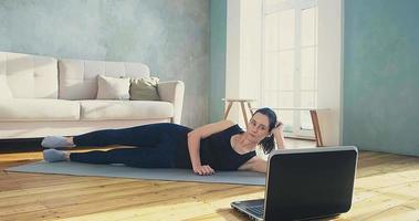 Athletic woman in stylish sportswear does supine side leg raises watching online training on laptop in living room slow motion video