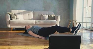 Brunette lady in stylish sportswear does crunches and leg lifts near laptop at online training in living room slow motion video