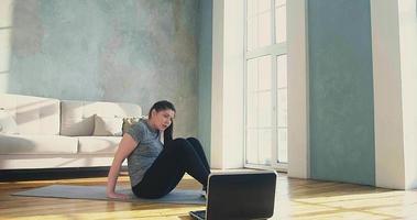 sportieve vrouw in stijlvolle sportkleding doet buikspieroefeningen met een laptop op een houten vloer bij online training in slow motion op de kamer video