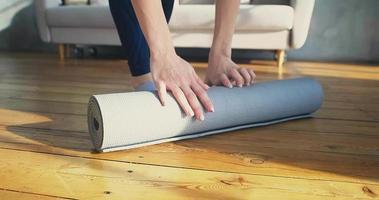 Young sportswoman in socks unrolls light grey rubber yoga mat on wooden floor in light room in morning closeup slow motion video