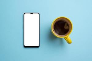 Phone mockup and coffee mug on clean blue surface. Top view, flat lay composition. Smart phone with blank, white isolated screen for app presentation