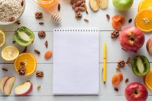 cuaderno con bolígrafo rodeado de manzanas, kiwi, frutos secos, naranjas y manzanas. el concepto de una dieta saludable y una lista de compras. foto