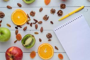 cuaderno con bolígrafo rodeado de manzanas, kiwi, frutos secos, naranjas y manzanas. el concepto de una dieta saludable y una lista de compras. foto