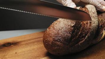 Woman in latex gloves cuts loaf of fresh rye bread with sharp knife on wooden cutting board on black background macro slow motion