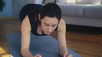 Concentrated calm woman in stylish tracksuit does plank exercise on grey mat near sofa in living room closeup slow motion